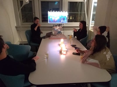 Team JUNGMUT sitzt im Konfi-Raum um den Tisch. Kerzen in der Mitte, Bildschirm zeigt eine Präsi.