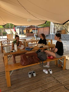 Drei Frauen sitzen unter Zeltdach auf Holzbänken und unterhalten sich.