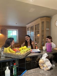 Drei Frauen sitzen an Küchentheke und lachen. Davor steht ein Obstkorb.