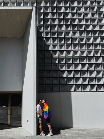 Zwei Männer mit Regenbogenflagge küssen sich vor grauem Betongeböude.