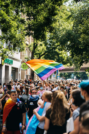Menschenmenge. Menschen im Vordergrund umarmen sich, in der Mitte große Regenbogen-Fahne.